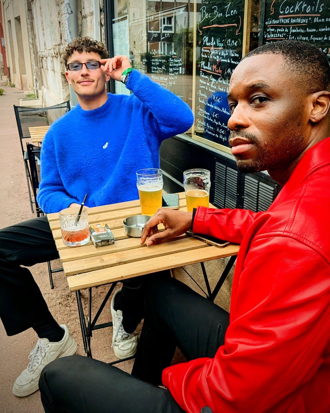 Deux amis en terrasse, pull bleu et veste rouge, bières sur la table